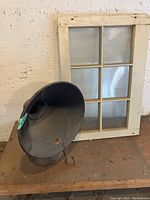 Front and side view of the black metal gramophone speaker with attached Atwater Kent label and small wooden 6-pane window behind it, showing dust and wear.