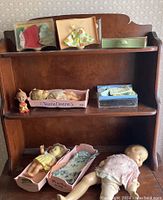 Vintage Tearie Dearie dolls and outfits arranged on a wooden shelf with boxed baby dolls, small figurine, doll outfits in packaging, and cradles.