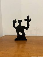 Front view showing abstract humanoid figure with two raised arms on a wooden surface