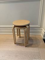 Two IKEA FROSTA molded plywood stools stacked, round seats visible, light wood floor and wall background