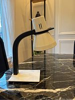 Photo of vintage postmodern desk lamp on a black marble table, showing side view with flexible black arm, white conical shade with ventilation slots, and white square base.