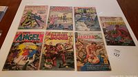 Seven vintage comic books laid out on table, titles include Star Spangled War Stories, Batman, Tarzan, Angel, Fightin' Scout, Dinosaurs. Covers show aging and rough condition.