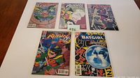 Five DC Comics comic books laid out flat on a wooden surface, 4 featuring Robin and 1 featuring Batgirl, all in protective sleeves.
