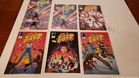 Front view of six comic books laid out on a white surface showing colorful covers with characters in superhero poses and energy blasts.