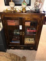 Front view of antique wood cabinet with etched glass doors, showing books inside and three bottom drawers with brass handles. Two lamps placed on top.