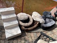 Four fabric dog beds and foam dog stairs on brick patio with orange safety fence in background.