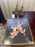 Photo shows two Thai metal figurines with turquoise and gold coloring and detailed headdresses on a wooden table beside a framed Thai painting with gold frame and colorful figures.