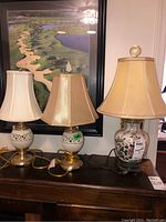 Three table lamps with various shades and ceramic bases displayed together in front of a golf course print on the wall.