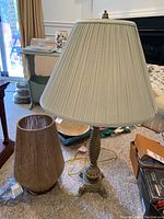 Lamp with carved wooden spiral base and fabric pleated lampshade beside lamp with resin base and string-wrapped shade on carpeted floor.