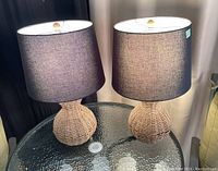 Photo showing two table lamps with woven bases and gray fabric shades on a glass table.
