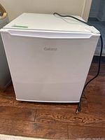 Front view of white Galanz mini fridge with power cord on wooden floor.