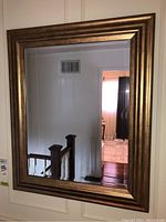 Mirror hung on wall above stair railing with beveled edge and gold frame reflecting hallway and door