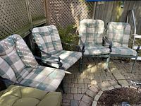 Four metal frame outdoor armchairs with plaid cushions arranged in outdoor space with stone tile flooring and wooden fence.