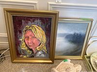 Two framed paintings leaning against a wall. Left: portrait of blonde woman with a reddish background signed by Guarek, showing canvas damage. Right: misty landscape painting with trees signed Piotrwoski 75.