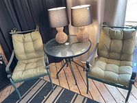 Photo showing two metal frame outdoor armchairs with tufted green cushions and a round glass-top metal frame table set inside a room on wood floor.