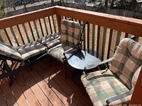 Photo of two metal frame armchairs with green and beige checkered fabric cushions alongside round wicker-style side table with glass top on wooden deck