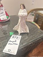 Porcelain figurine of a woman in white winter cloak holding a muff, standing 6 inches tall, displayed on a wicker table with a maxsold tag indicating 'Royal Doulton'.