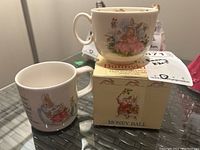Two Royal Doulton Bunnykins mugs and money ball, one on top of money ball's original box showing Bunnykins logo and packaging