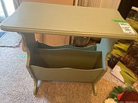 Angled view of vintage sage green painted wooden side table showing one magazine rack with scalloped edges beneath the flat top surface.