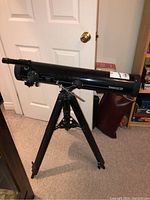 Full view of the black Tasco telescope mounted on an adjustable tripod inside a room with beige carpet.