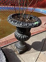 Front angle view of black resin plant urn outdoors by red plastic fencing, showing soil with plant stems inside and urn base wear.