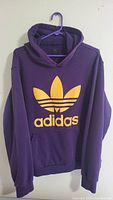 Purple Adidas hoodie with large yellow Trefoil logo and text on the front, hanging on a hanger.