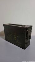 View of the entire rectangular metal ammunition box with handle on top and military stencil markings on the front side.