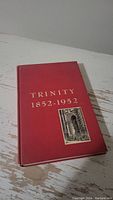 Closed hardcover book with red cover and gold embossed text 'TRINITY 1852-1952' with an attached small black and white image.