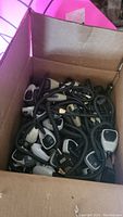 Box filled with numerous Motorola handheld radio microphones with coiled cables inside a cardboard box.