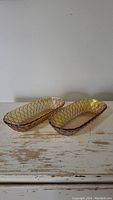 Two carnival glass oval trays with gold iridescent amber finish, pretzel pattern, placed on a rustic white wooden surface.