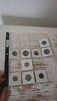 Photo showing nine coins in white cardboard holders, some labeled with countries and years mounted in a clear plastic sheet. Coins show varying degrees of wear.