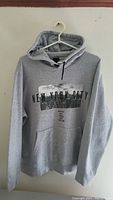 Full view of grey hoodie with New York City graphic on front hanging on white plastic hanger against wall.