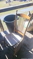 Wooden rocking chair with curved rockers and spindle backrest, has a light-colored seat cushion, shown with sunlight casting shadows, located next to a large black trash bin outdoors.