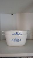 Two stacked white Corning Ware casserole dishes with blue cornflower design on shelves.