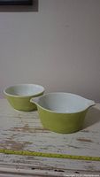 Two light green Pyrex mixing bowls with white interiors on a distressed wooden surface. One bowl has no spouts, the other has dual pour spouts.