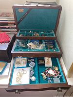 Wooden jewelry box open showing two-tier design, top tray and lower drawer lined in teal velvet with assorted jewelry.