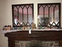 Front view of the miniature Christmas village arranged on a wooden mantle with two large mirrors behind it. Several buildings and figurines visible with warm interior lighting in the houses.
