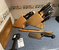 Two wooden knife blocks on kitchen counter with various knives, rustic wooden bread cutter, cleaver, honing steel, and sharpener in view