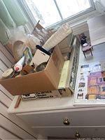 View of project lot including cardboard box with crafting supplies, dollhouse kit box, and the dolls house book on a dresser under a window.