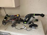 Both Dyson V7 Trigger and Bissell MultiReach handheld vacuums with wands, attachments, and charger on table surface