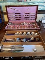 Photo showing multiple utensils stored in wooden trays and a pink lined box, including forks, spoons, knives, and serving utensils.