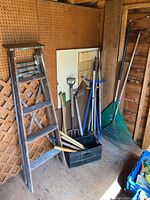 Overall view of lot showing wooden step ladder, plastic leaf rakes, metal garden rake, and black crate containing smaller tools.