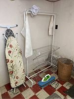 Overall shot showing two ironing boards, white metal drying rack, green and white electric iron with blue iron rest, and round wicker basket on basement floor with red and white tile.