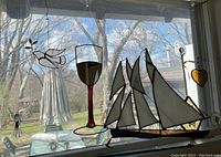 Photo showing stained glass ornaments of a dove, wine goblet, sailboat, and small amber heart hanging on window with trees outside