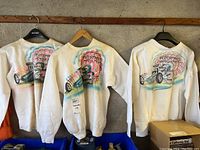 Three white crewneck sweatshirts hung on hangers showing back airbrush designs