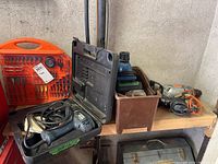 Photo showing drills, cases, drill bits set in orange case, and various tools on wooden garage shelf.