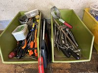Two green plastic bins filled with assorted hand tools including screwdrivers, pliers, hammers, and wrenches on a workbench.