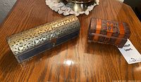 Photo showing both trinket boxes on wooden table; larger metal and wood box with cut-out pattern and smaller carved wooden box.