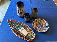 Five artisan pottery pieces arranged on blue covering: two vases, one creamer jug, two decorative plates including elongated dish with colorful abstract glaze