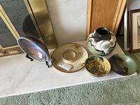 Display of five handcrafted pottery items on a white surface near a fireplace: a large iridescent bowl on a display stand, a beige terracotta dish, a decorative stoneware vase, and two smaller artistically glazed bowls.